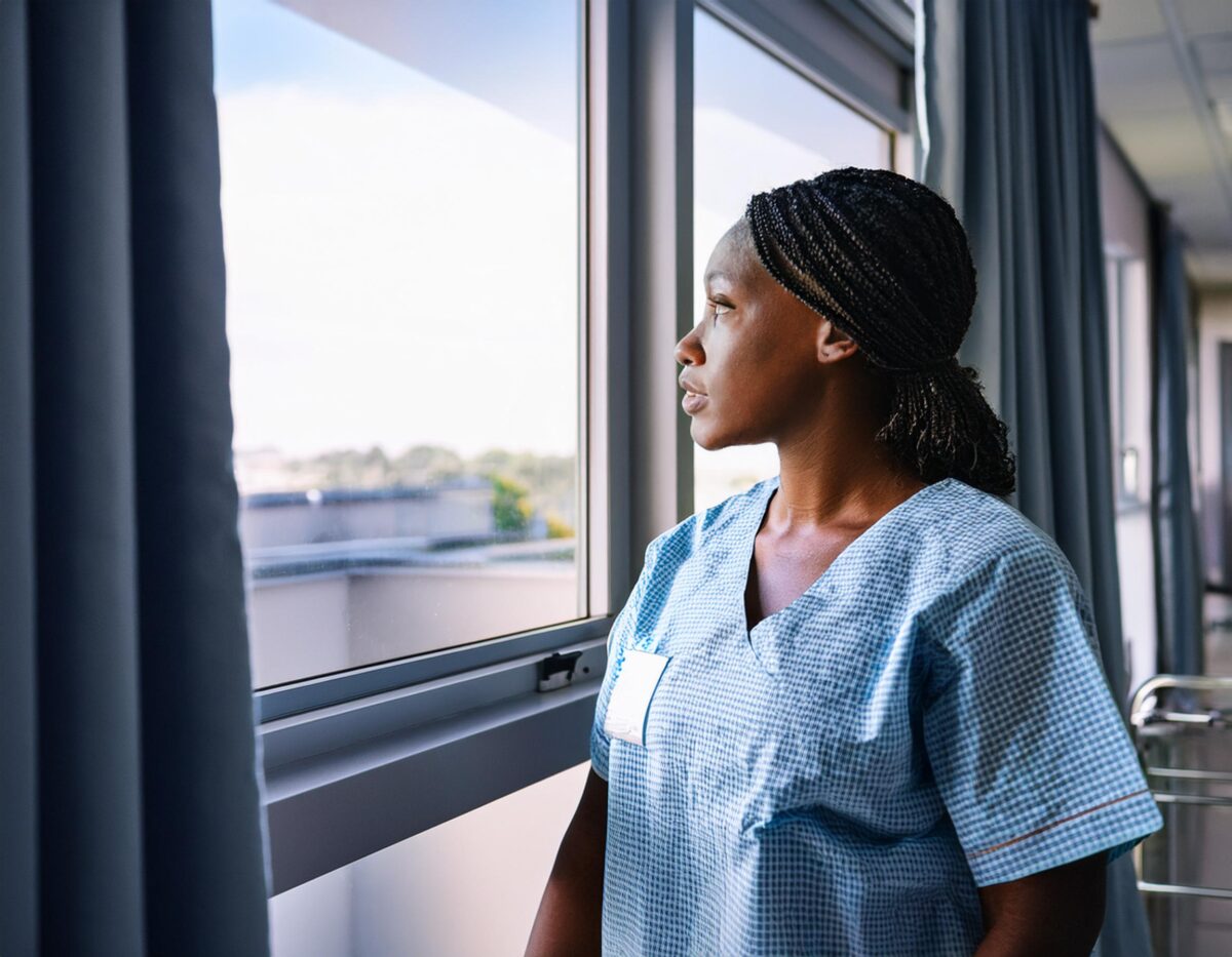 A woman in a hospital gown sitting on a bed, gazing out the window.