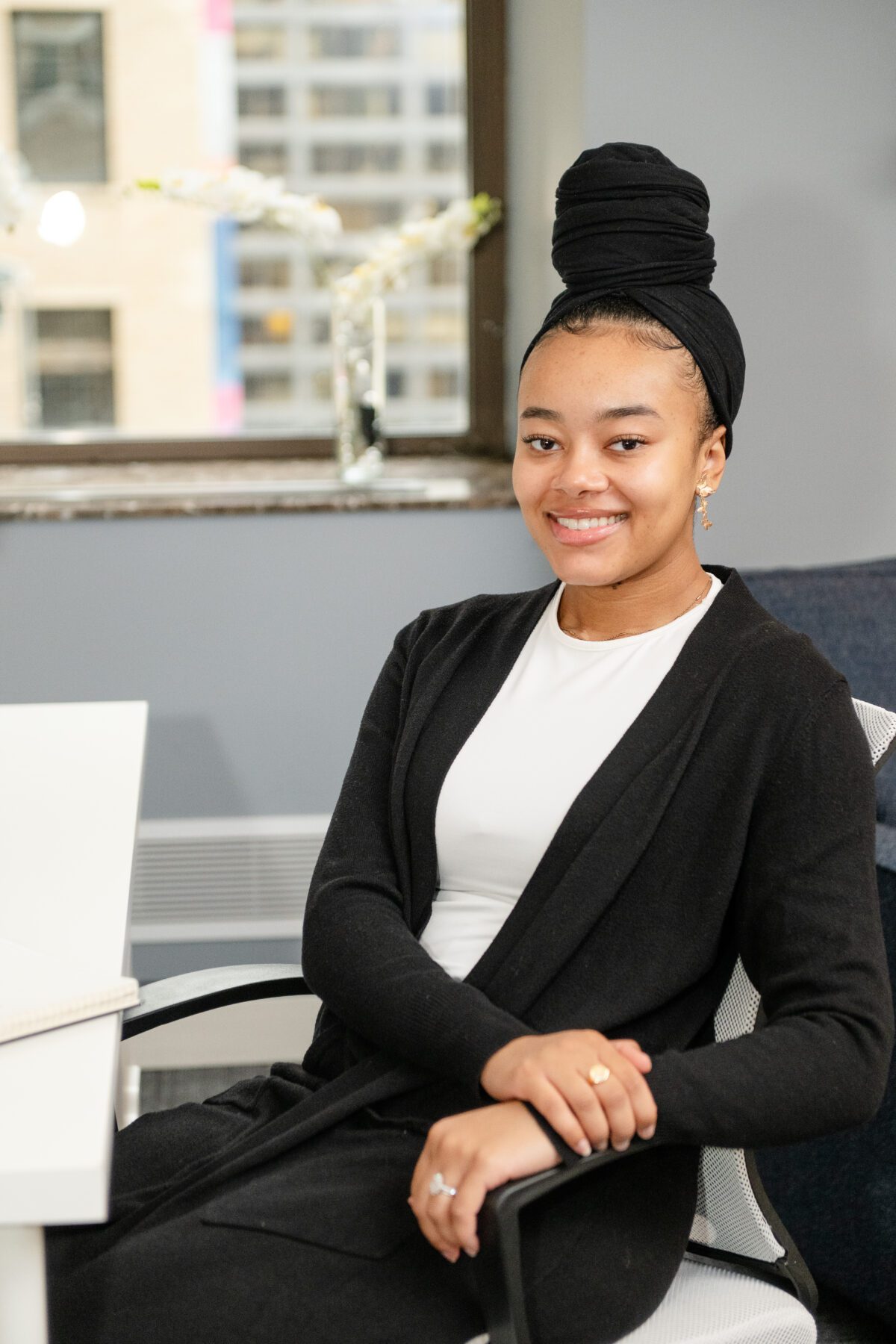 Kalila F., an intern therapist, sits confidently in an office chair, smiling. She wears a black cardigan over a white top with a black headscarf, framed by a window with natural light.